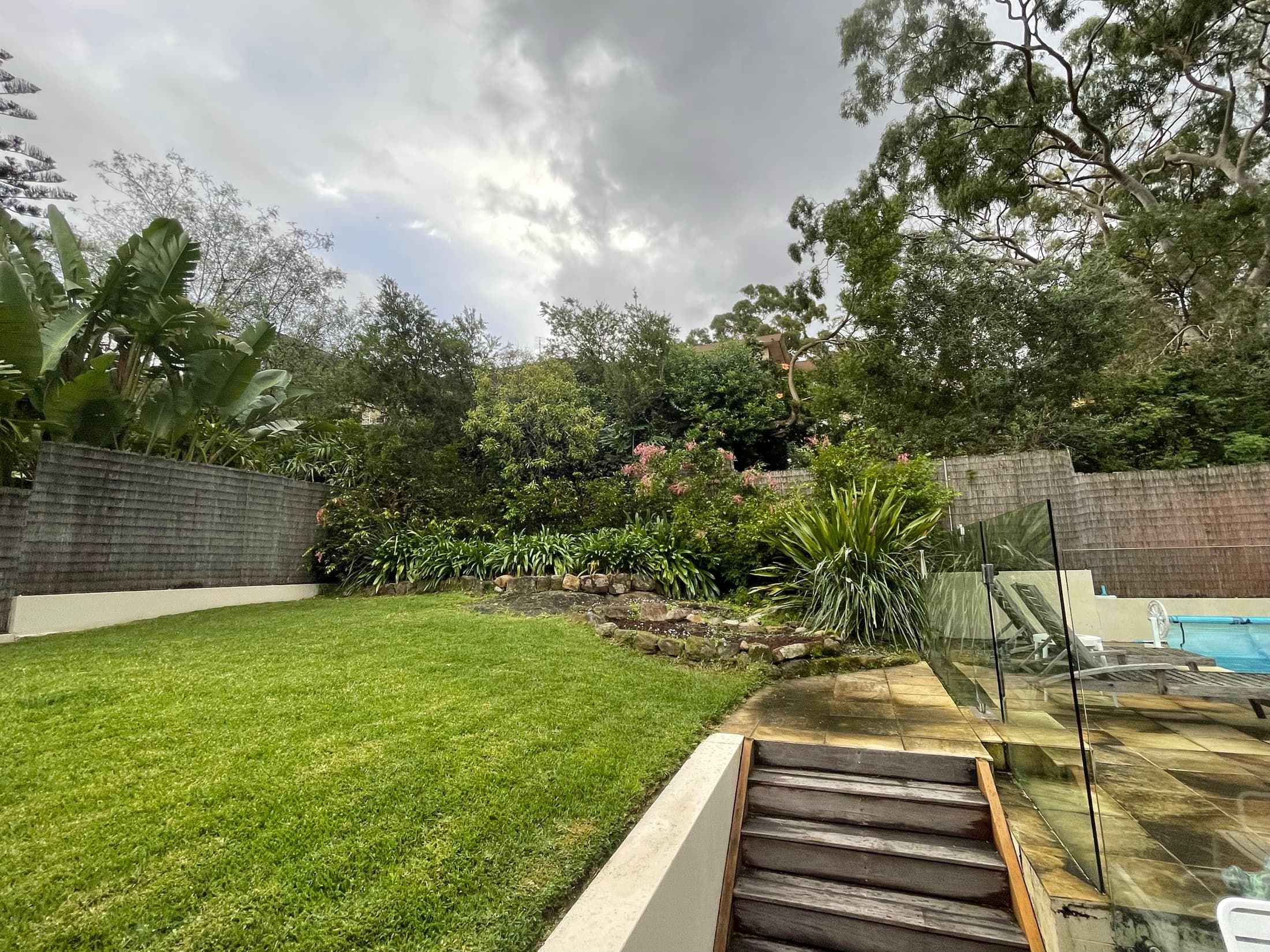 The rear yard before the works, lawn running to a rubble edge and an ageing timber pool surround